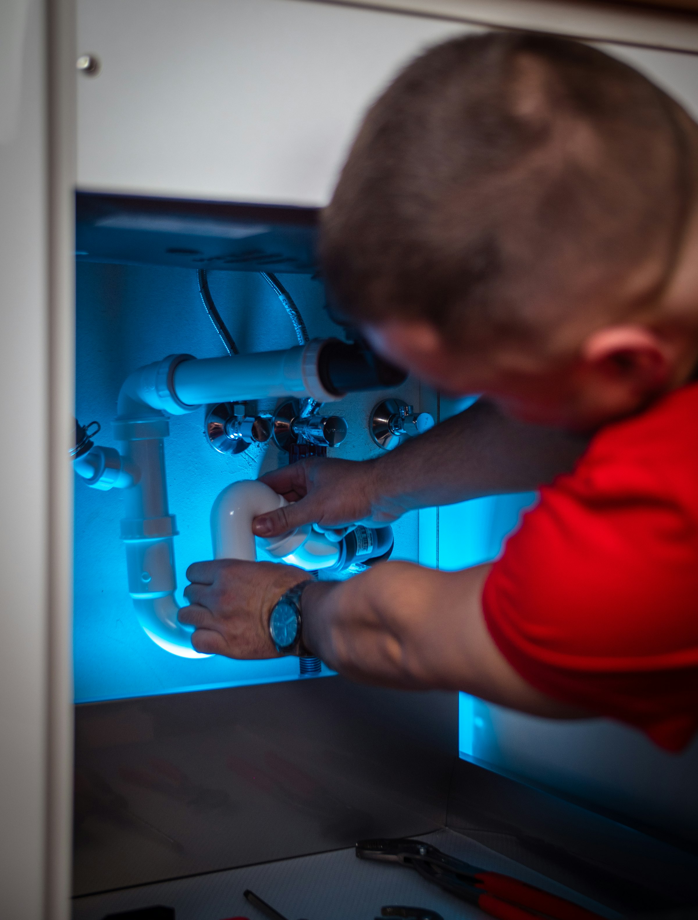 A plumber repairing pipework under a sink, lit by a work light